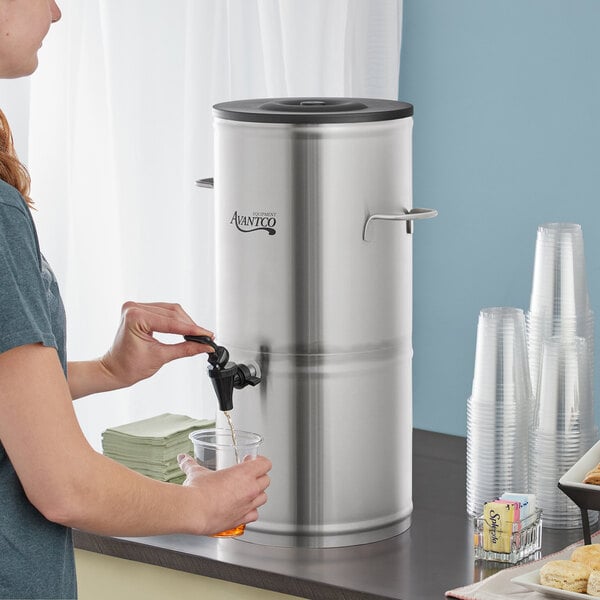 A woman pouring water into an Avantco stainless steel iced tea dispenser.
