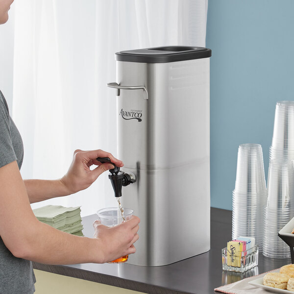 A woman using a stainless steel faucet to pour liquid from an Avantco slim iced tea dispenser.