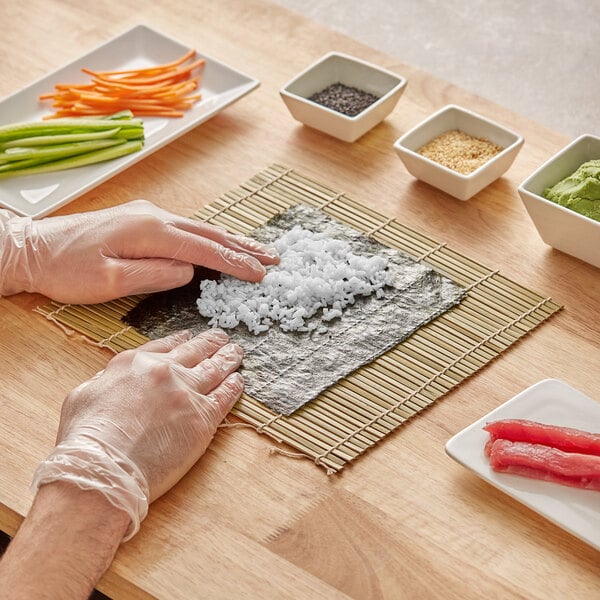 A person in plastic gloves using a bamboo sushi rolling mat to make sushi on a table.