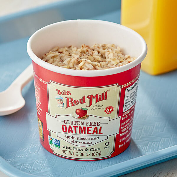 A container of Bob's Red Mill Apple Cinnamon oatmeal with a label on a table with a bowl of oatmeal and a spoon.