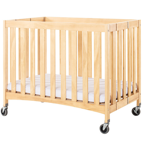 A natural wood slatted folding crib with wheels and a white sheet.