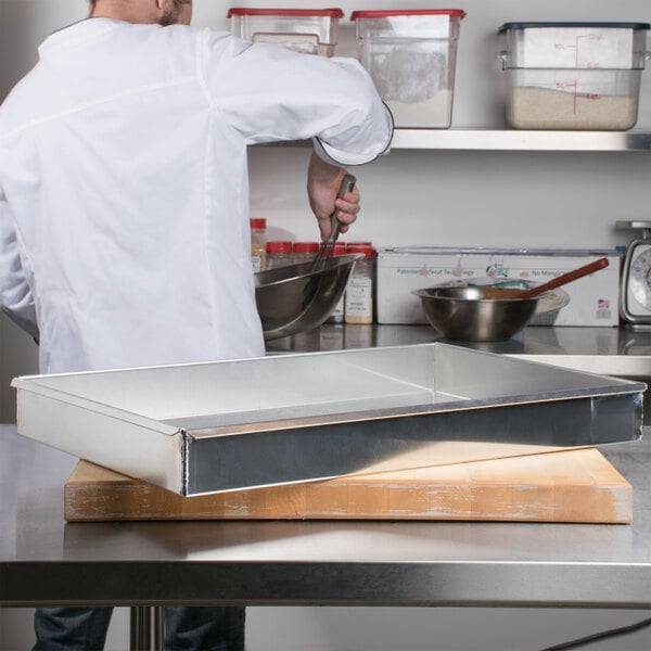 A man in a white coat mixing food in a Vollrath aluminum cake pan on a metal surface.