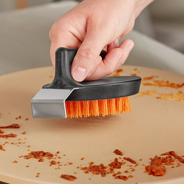 A hand using an Outset Pizza Stone cleaning brush with a scraper to clean a pizza stone.