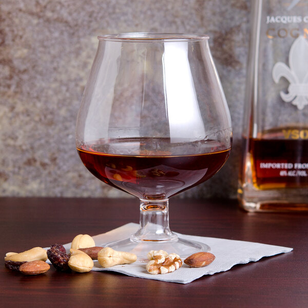 A customizable plastic brandy snifter filled with brown liquid on a table with nuts and a bottle of liquor.