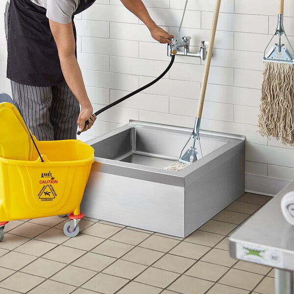A man using a yellow bucket to clean a Regency stainless steel mop sink.
