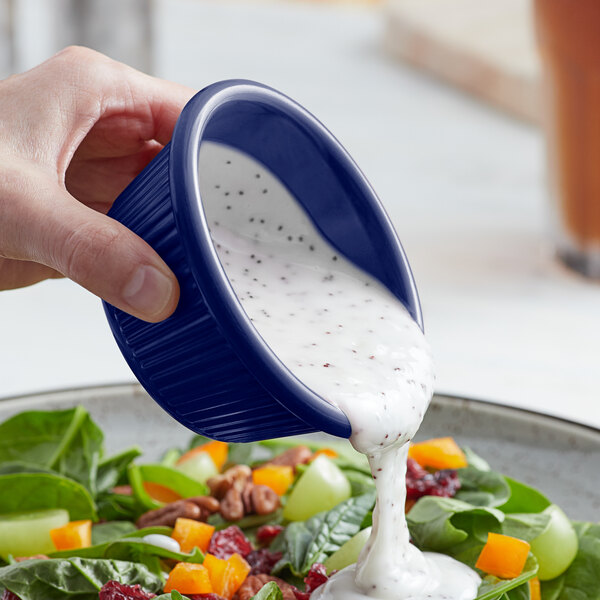 A person using a blue Acopa fluted ramekin to pour white dressing onto a bowl of salad.