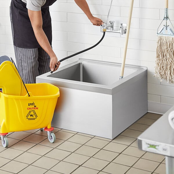 A man cleaning a white Regency stainless steel mop sink with a yellow mop bucket.
