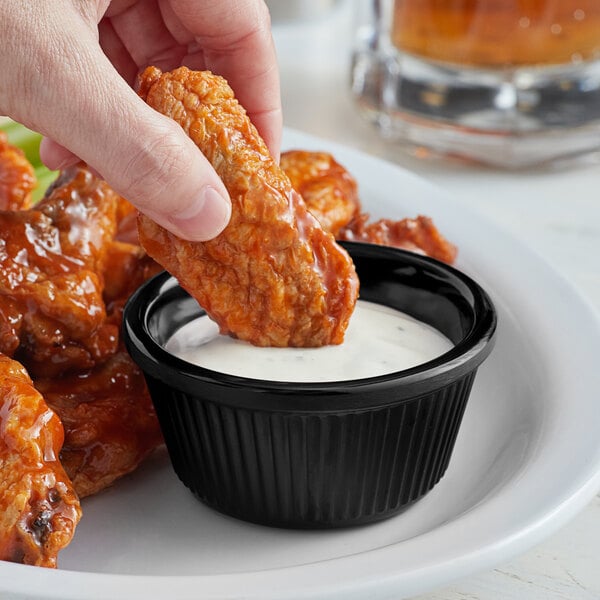 A person dipping a chicken wing into a black Acopa fluted ramekin of sauce.