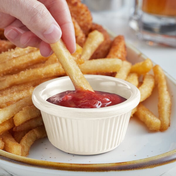 A hand dipping a french fry into an Acopa Bone White fluted ramekin of ketchup.