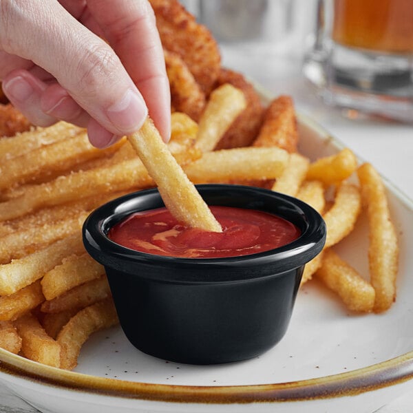 A hand holding a french fry dipping it into a black Acopa ramekin full of ketchup.