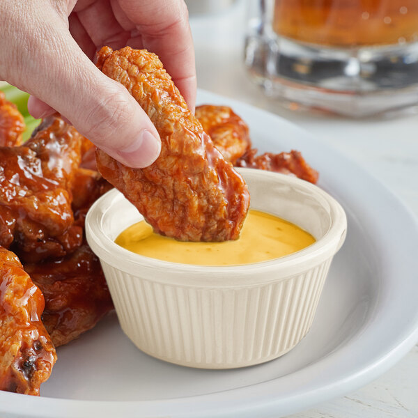 A hand dipping a piece of chicken into a white fluted Acopa ramekin of sauce.