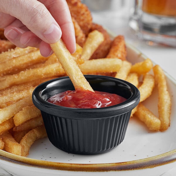 A hand holding a french fry dipping it into a black Acopa fluted ramekin of ketchup.