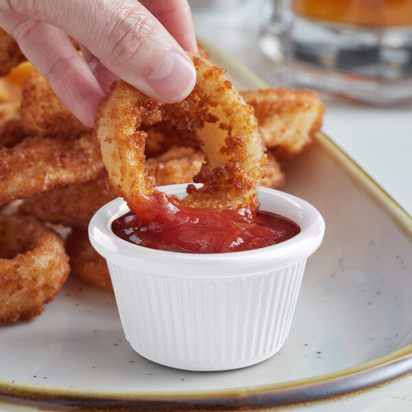 A hand dipping a fried onion ring into a small white Acopa ramekin of ketchup.