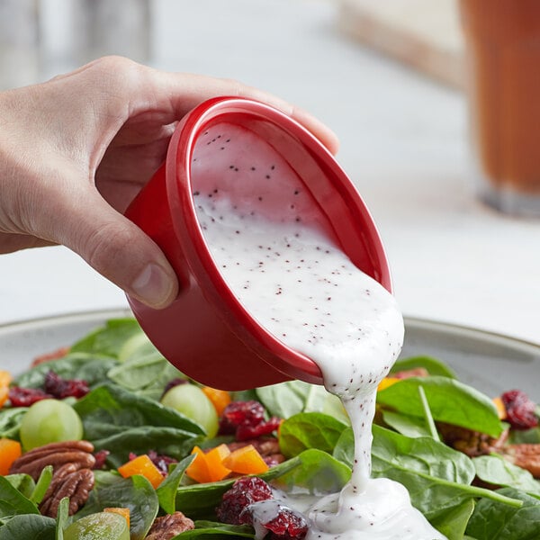 A hand pouring white dressing into a red Acopa ramekin.