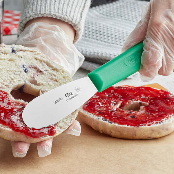 A person using a Choice stainless steel sandwich spreader with a green handle to spread jam on a bagel.