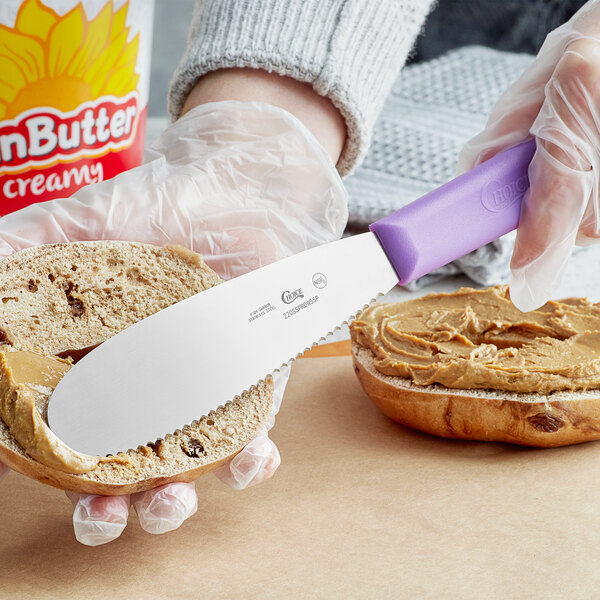 A person using a Choice stainless steel sandwich spreader with a purple allergen-free handle to spread peanut butter on a piece of bread.