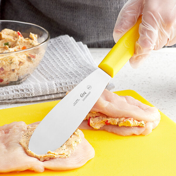 A person using a Choice Stainless Steel Sandwich Spreader with a yellow handle to spread butter on a chicken breast.
