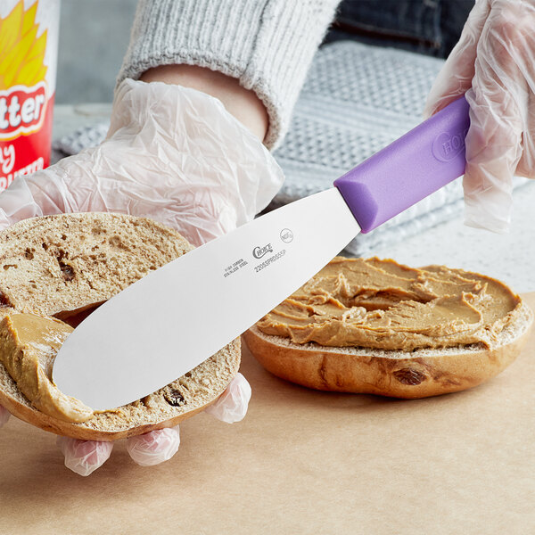 A person spreading peanut butter on a piece of bread using a Choice stainless steel sandwich spreader with a purple handle.