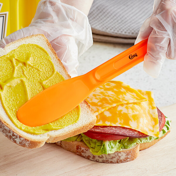 A gloved hand uses a Choice neon orange sandwich spreader to spread butter on a sandwich.