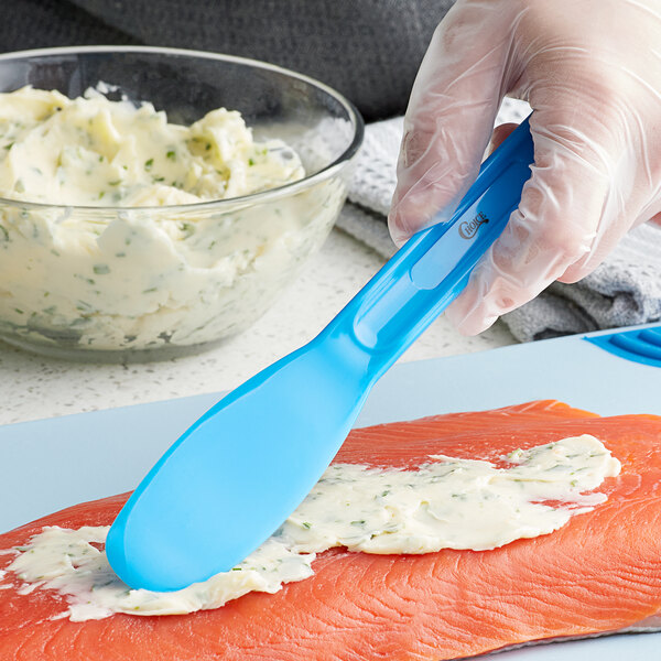 A person's hand with a Choice blue polypropylene sandwich spreader spreading cream on a piece of meat.