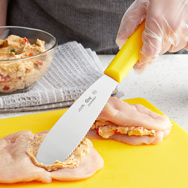A hand in gloves holding a Choice yellow scalloped sandwich spreader cutting a chicken breast.