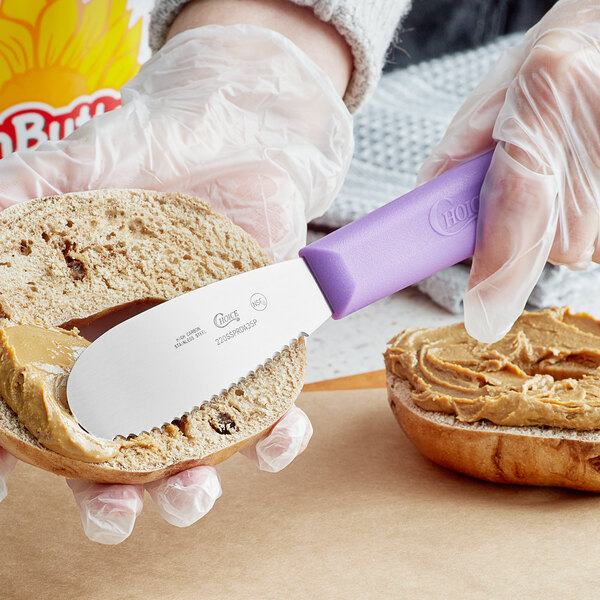 A person with a gloved hand using a purple Choice sandwich spreader to spread peanut butter on bread.