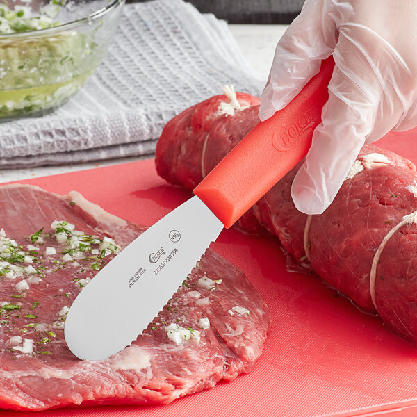 A gloved hand using a Choice stainless steel scalloped sandwich spreader to cut meat.