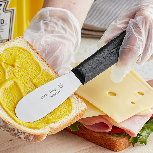 A person holding a Choice stainless steel sandwich spreader with a black handle and spreading butter on a sandwich.