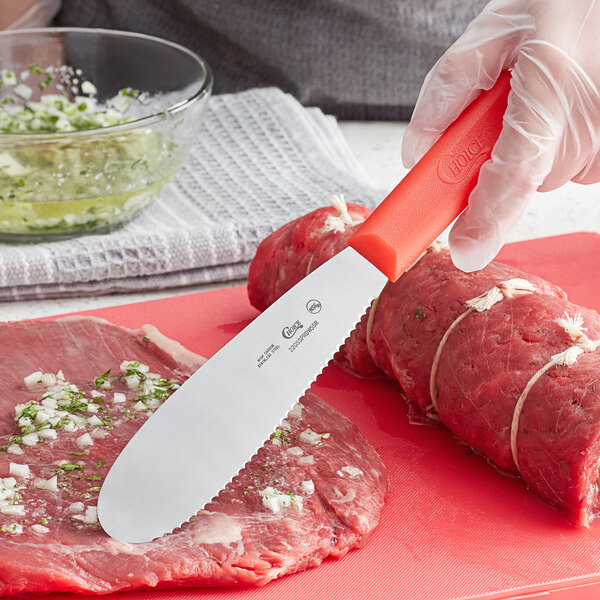 A person using a Choice stainless steel sandwich spreader with a red handle to cut meat on a red cutting board.