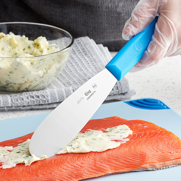 A person's hand using a Choice stainless steel sandwich spreader with a blue polypropylene handle to spread butter on a piece of fish.