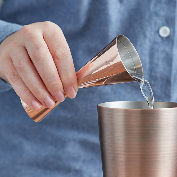 A hand pouring liquid into a copper-plated Barfly Slim Style Jigger.