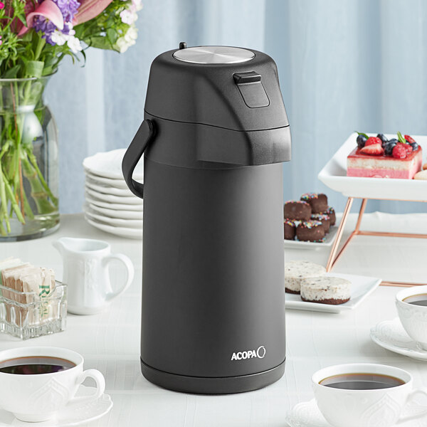 An Acopa matte black stainless steel airpot on a table with cups of coffee and desserts.