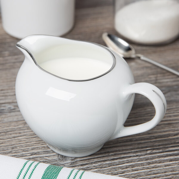 A white porcelain creamer with a silver handle on a table with a spoon.