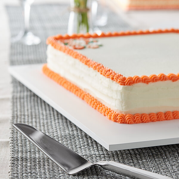 A square cake with orange icing on a white Melamine-coated wood cake board.