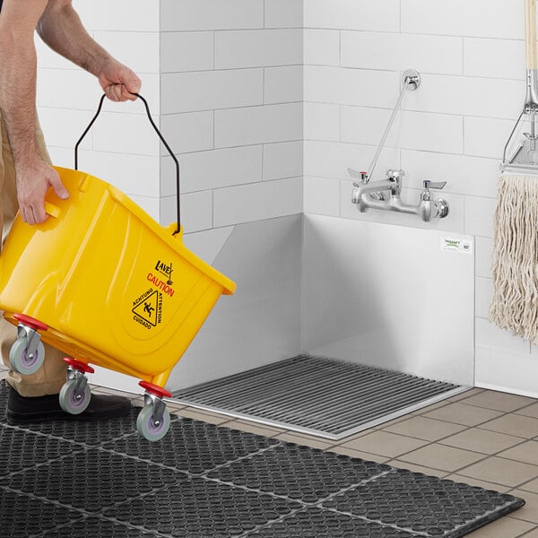 A person pushing a yellow bucket under a Regency stainless steel mop sink.