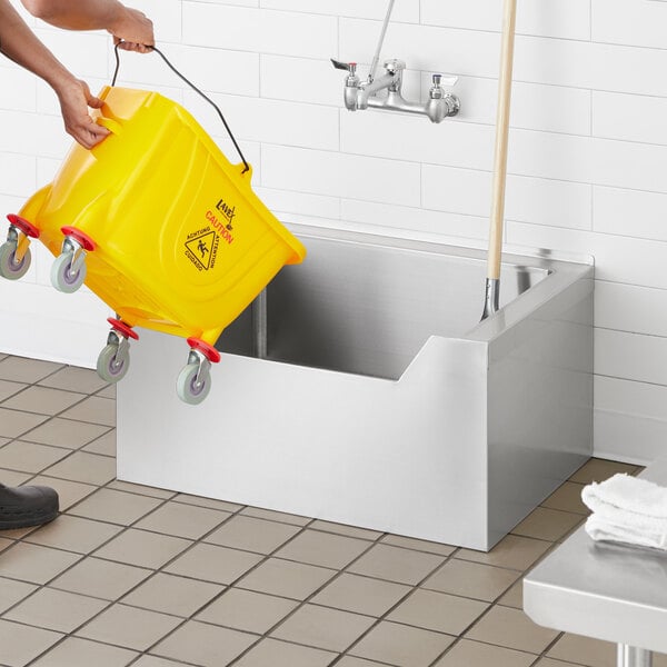 A person using a yellow bucket to fill a Regency stainless steel floor mop sink.