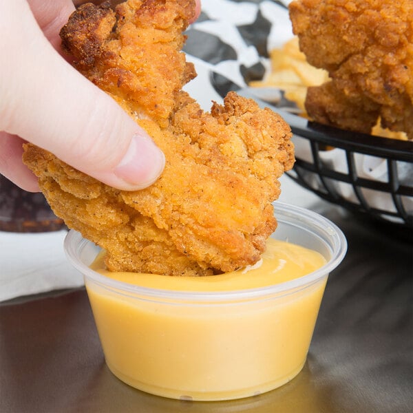 A hand holding a piece of fried chicken over a Dart clear plastic souffle cup of sauce.