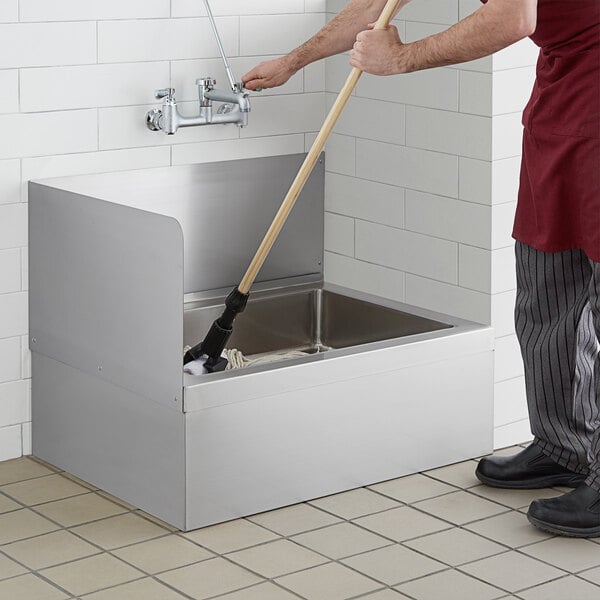 A person cleaning a Regency stainless steel mop sink with a mop.