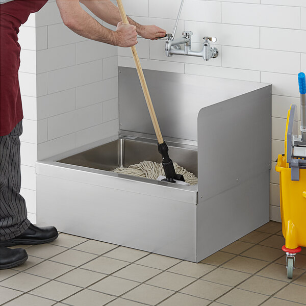 A person cleaning a stainless steel mop sink with a mop.