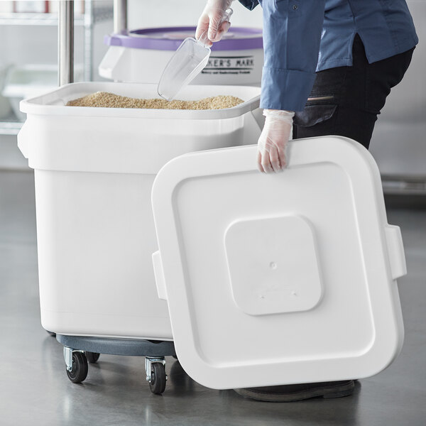 A person in a blue shirt and gloves putting brown rice into a white Baker's Mark ingredient storage bin.