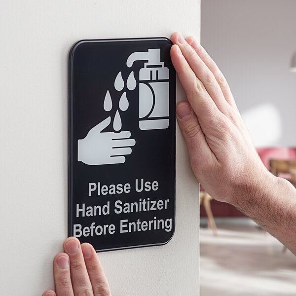 A person's hands holding a black and white plastic sign that reads "Please use hand sanitizer before entering" with a hand sanitizer symbol.