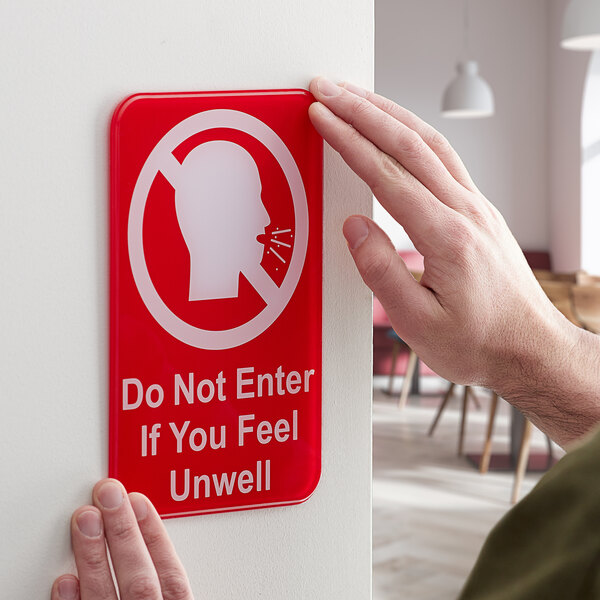 A person holding a red and white Tablecraft plastic sign with a head and text reading "Do Not Enter If You Feel Unwell"