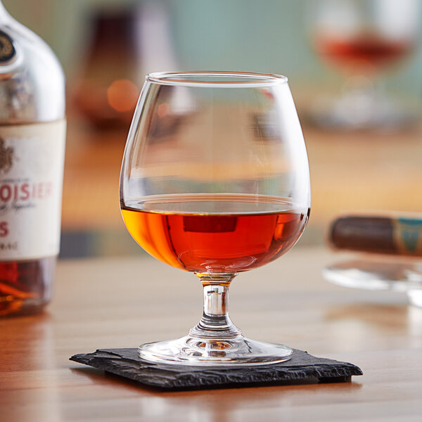 An Acopa Select brandy snifter filled with brown liquid on a coaster on a table.