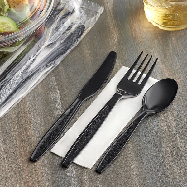 A black Visions plastic fork, spoon, and knife on a napkin next to a salad.