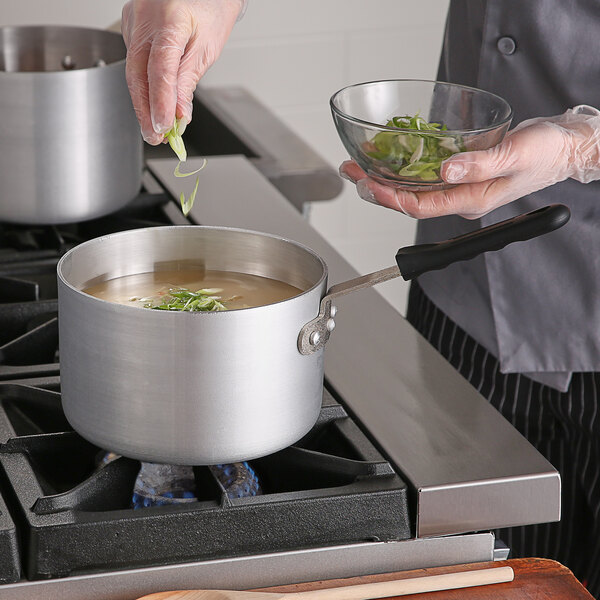 A person cooking soup in a Choice aluminum sauce pan on a stove.