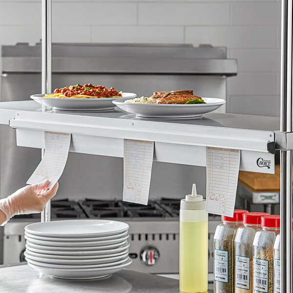 A woman in a professional kitchen holding a receipt over plates of food using a Choice Aluminum Wall Mounted Ticket Holder.
