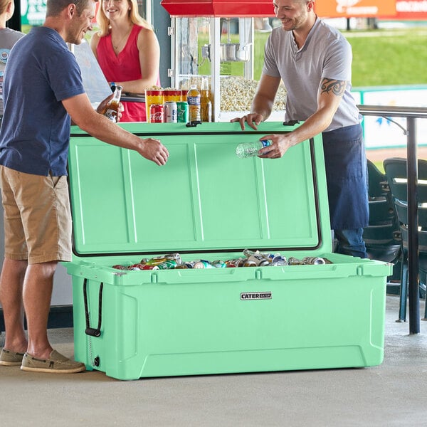 Two men standing next to a CaterGator Seafoam outdoor cooler with the lid open and a bottle of soda inside.