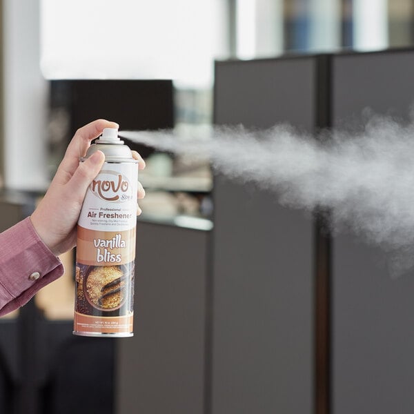 A person using a can of Noble Chemical Novo Vanilla air freshener to spray a room in a corporate office.