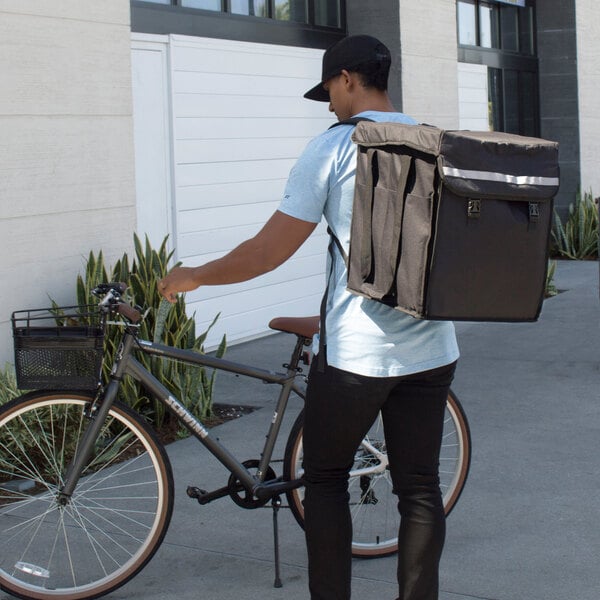 A man with a Cambro insulated delivery backpack on his back standing next to a bicycle.