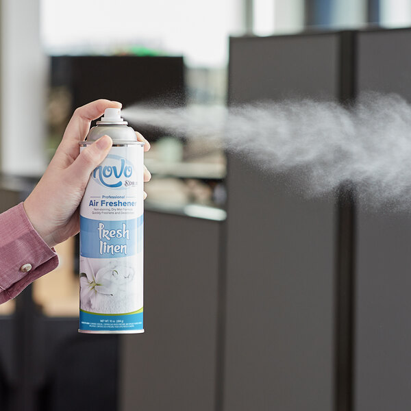 A person using a white and blue can of Noble Chemical Fresh Linen air freshener to spray air.